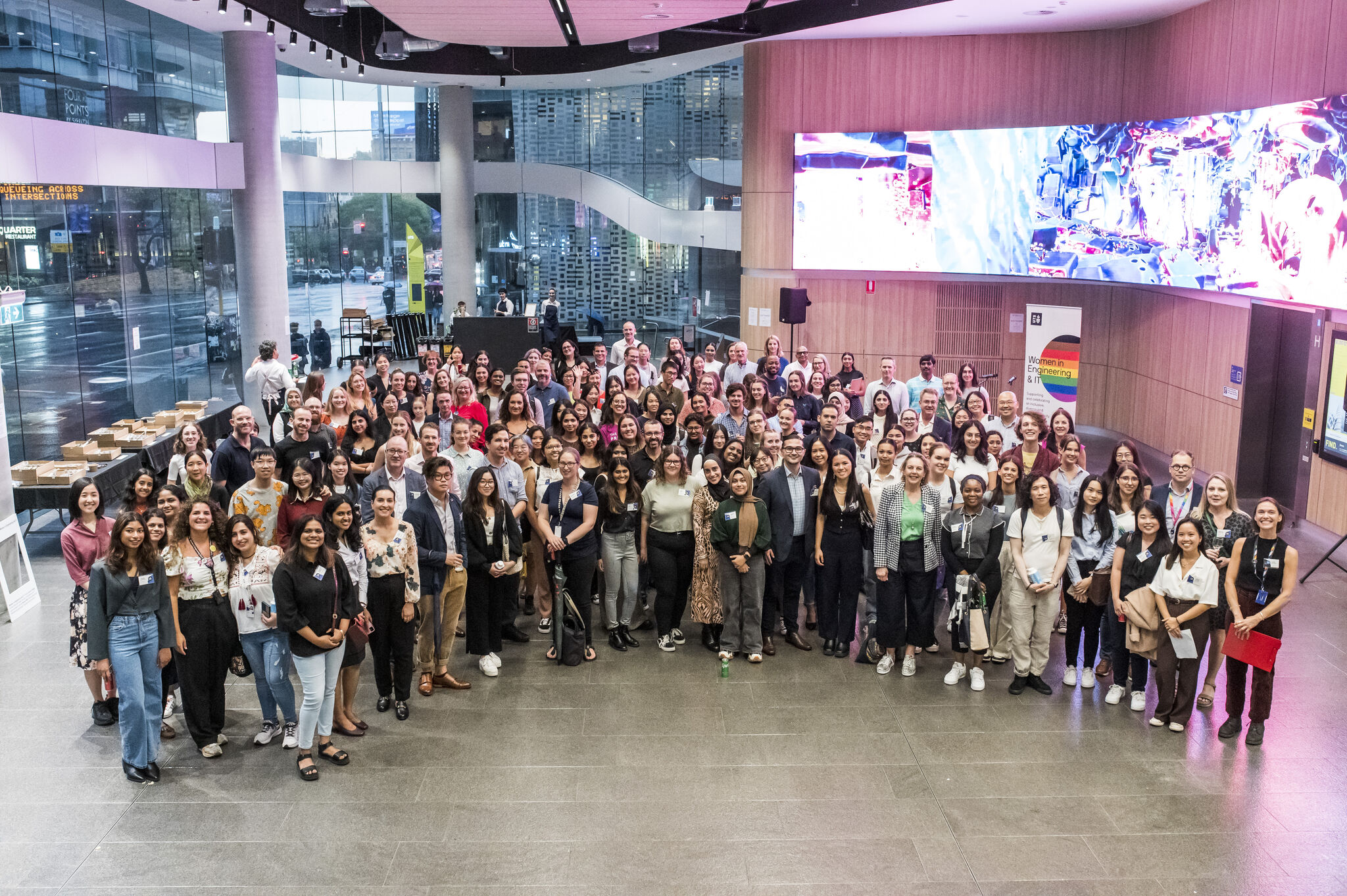 Large group at a Women in Engineering and IT gathering in a modern atrium with programme banner.